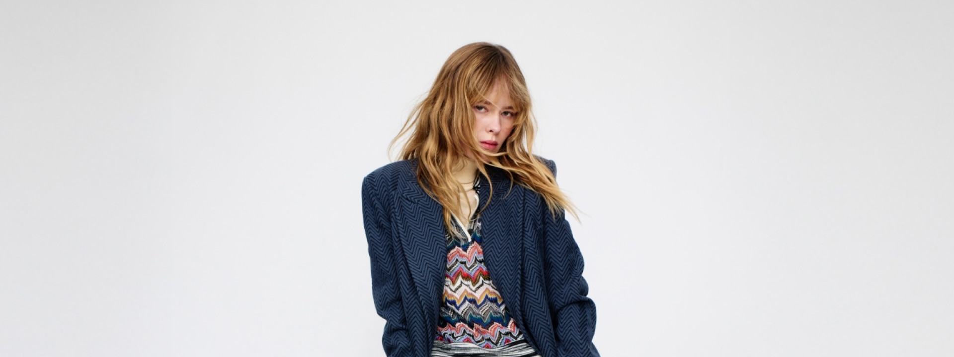 A model is wearing a blue suit and a zig zag multicoloured jumper