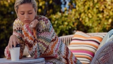 A model is sitting wearing a long dress with, behind here, different pillows in zig zag multicoloured texture