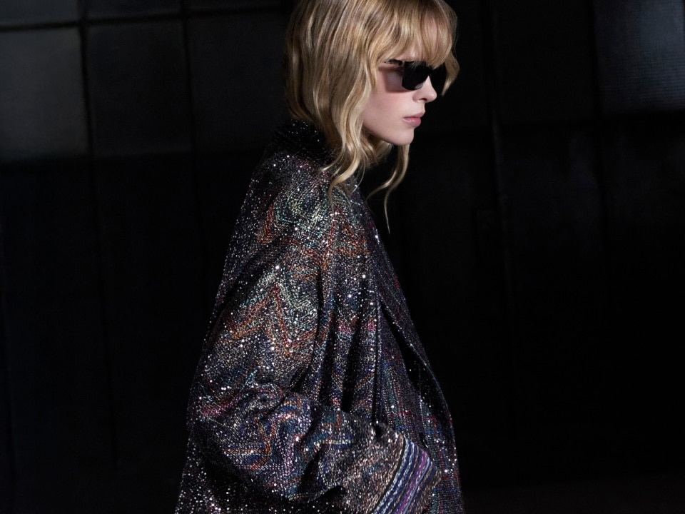 A model is wearing a long sleeveless dress with black, silver, and beige sequined striped pattern.