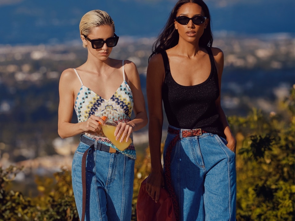Two models are wearing different outfits from the new SS25 colleciton: the first one has a light top, the second one a black  tank top, whikle both wearing denim